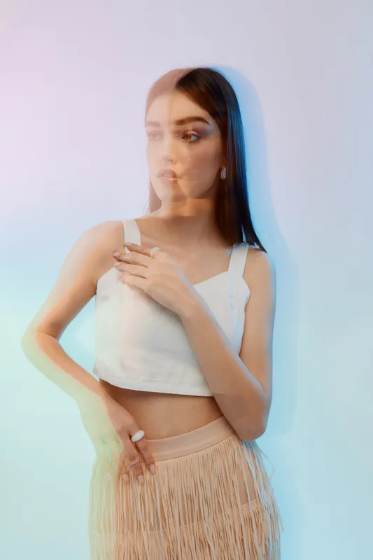 Young woman with long hair in white top and beige skirt poses with hand on chest. Soft colorful lighting creates artistic double exposure effect. Concept of beauty fashion and style.