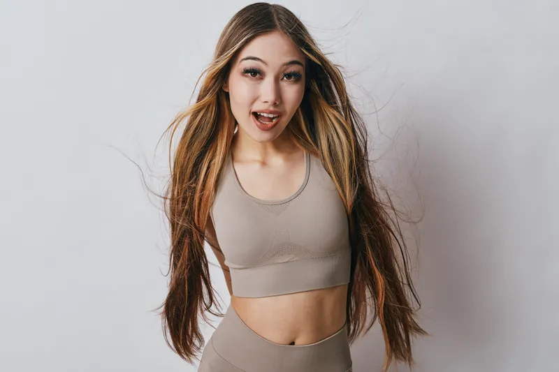 Young woman with long hair in sportswear looking surprised and happy with loose hairstyle against white background in studio. Fitness girl wearing beige activewear smiling indoors.