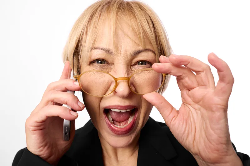 Elderly woman in black suit holding phone and glasses, screaming with panic and stress, anger visible, communication crisis intense emotion on clean white background