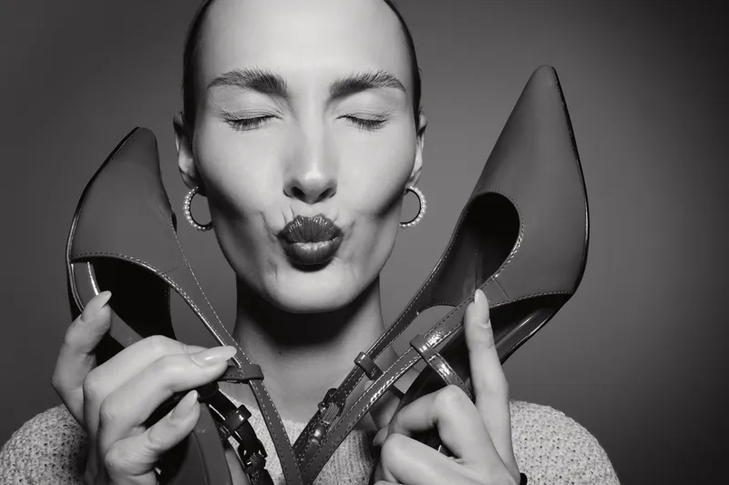 Close-up of a Caucasian woman with light skin holding high heels near face, eyes closed and lips puckered in a playful pose, studio lighting.