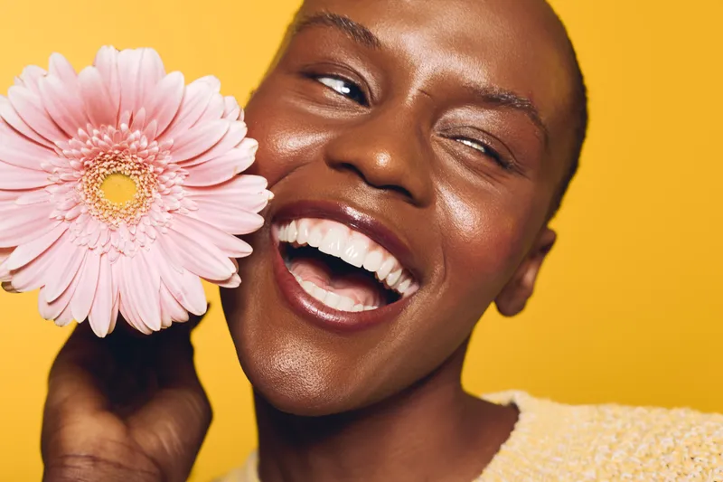 Happy African woman with dark skin laughing and holding a pink flower near her face on yellow background. Concept of natural beauty, joy, and floral wellness in studio portrait.
