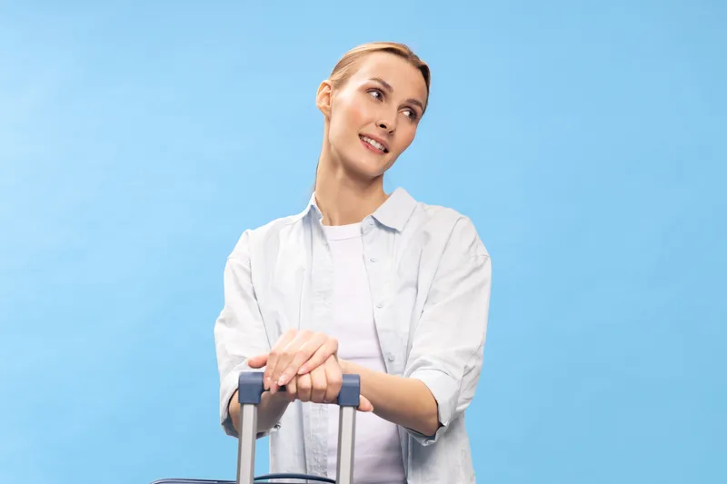 Young woman standing with luggage, looking sideways with a smile against blue background. Concept of travel, vacation, tourism, and casual lifestyle for young adults.