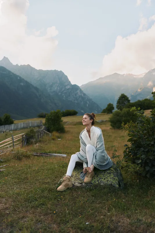 Young woman enjoys outdoor lifestyle in a mountain meadow, sits on a rock, wearing light cardigan and boots, tranquil landscape behind, calm mood and natural surroundings