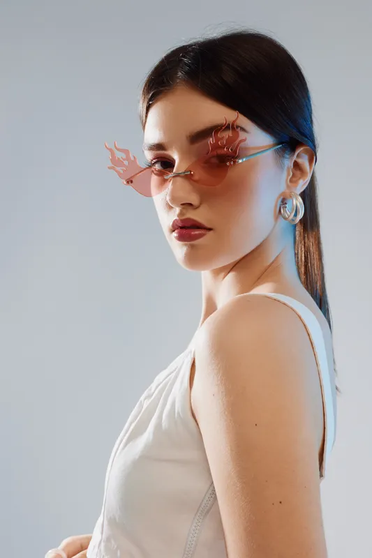 Young woman with sleek dark hair wearing unique pink sunglasses and silver hoop earrings poses confidently in a white sleeveless top. Concept of beauty fashion style.