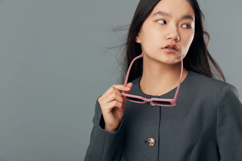 Young woman holding pink eyeglasses with hand, wearing grey jacket and looking to side on plain background. Female model with long dark hair posing for fashion or portrait studio shot.