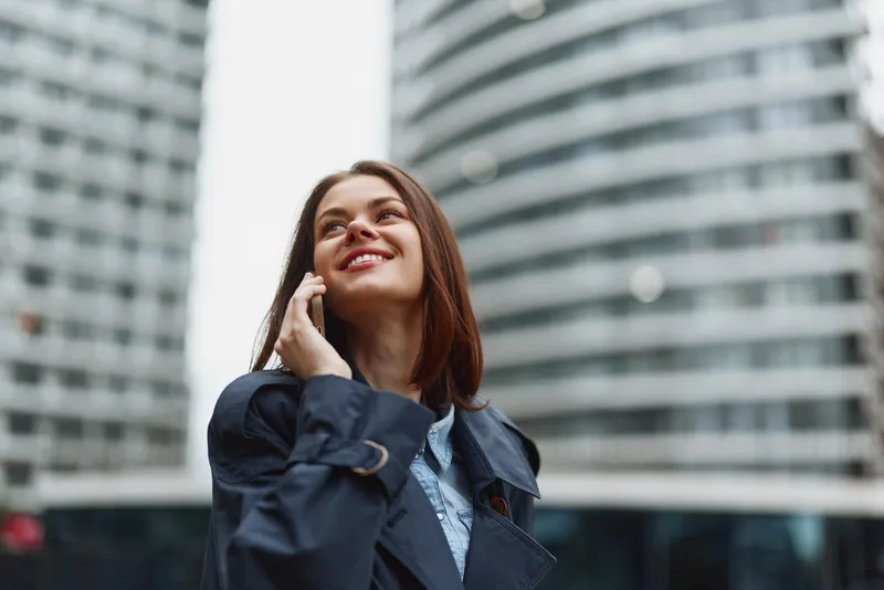 Confident woman outdoors in a navy coat chats on her phone, looking up with a bright smile among urban glass towers, capturing a candid moment