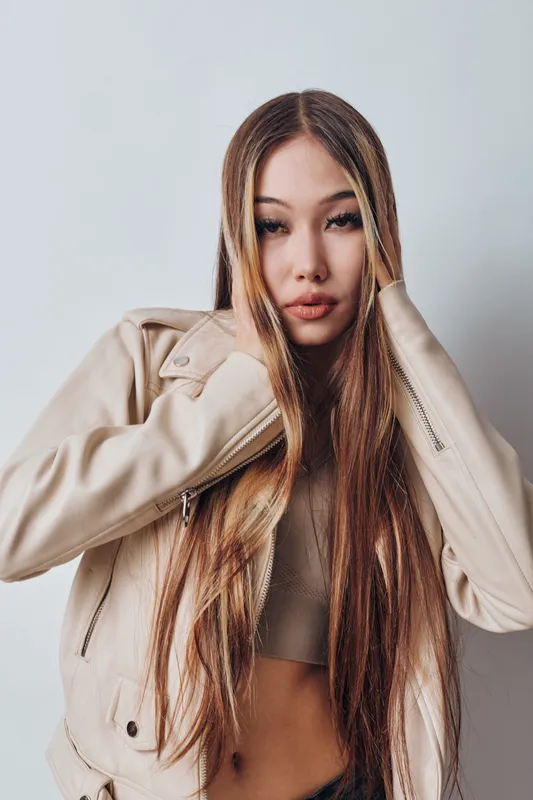 Young woman with long straight hair wearing beige jacket and crop top posing indoors against plain background for fashion and style portrait.