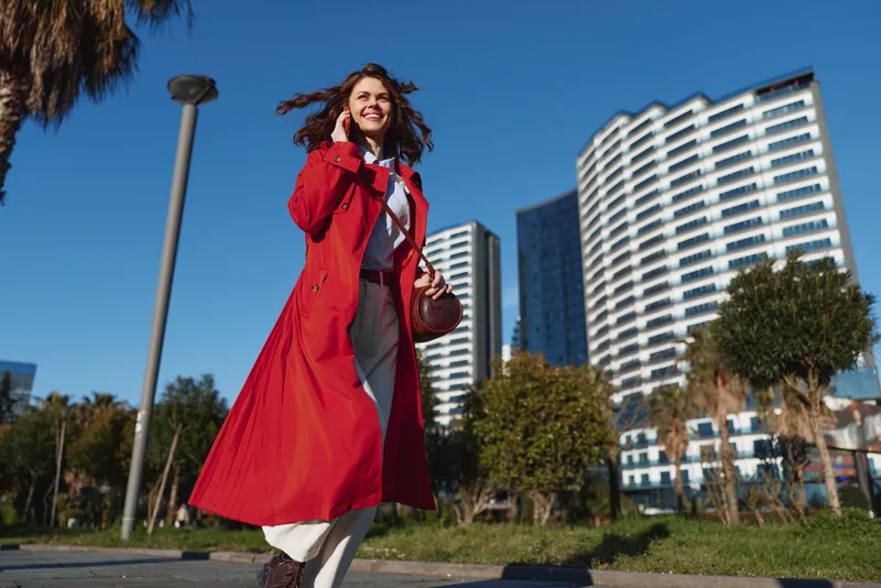 Young woman in a flowing red coat walks outdoors in an urban park while talking on her phone with a joyful expression on her face.