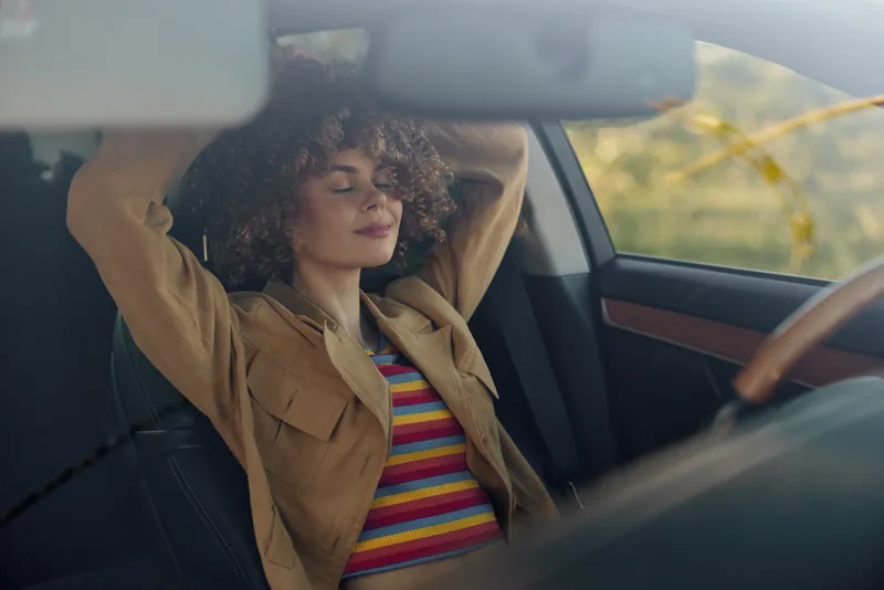 Relaxed woman in a vintage car rests with eyes closed, striped top and tan jacket, conveying casual lifestyle and warm retro film color.