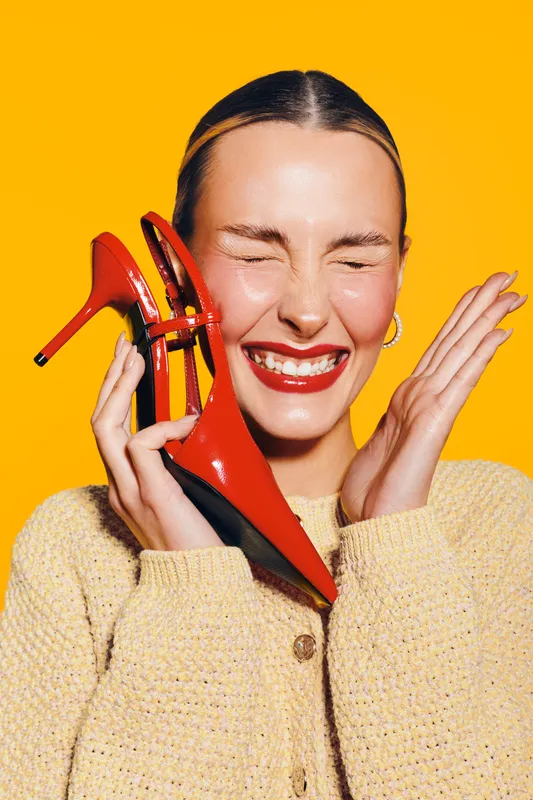 Happy woman with red lipstick holds red high heel shoe near face, eyes closed and smiling. Bright yellow background and cozy beige sweater create cheerful mood and fashion concept.