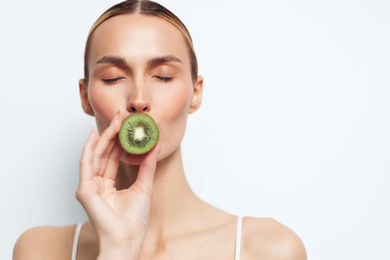 A young woman with blonde hair wearing a white top bites into a kiwi, eyes closed in contentment, savoring the fresh fruit in a bright minimalist studio with clean white walls