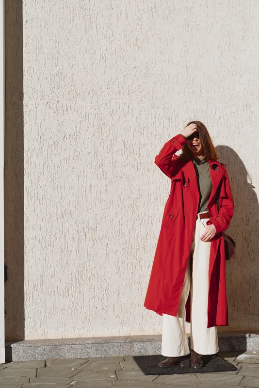 A stylish woman stands outdoors against a textured wall, wearing a bright red trench over a gray sweater and white wide-leg pants, shielding her eyes from bright sun with one hand.