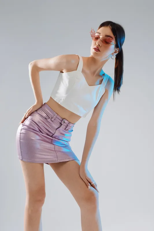 Young woman in a white crop top and shiny purple skirt poses wearing pink sunglasses. Fashion concept with beauty and style on a neutral background.