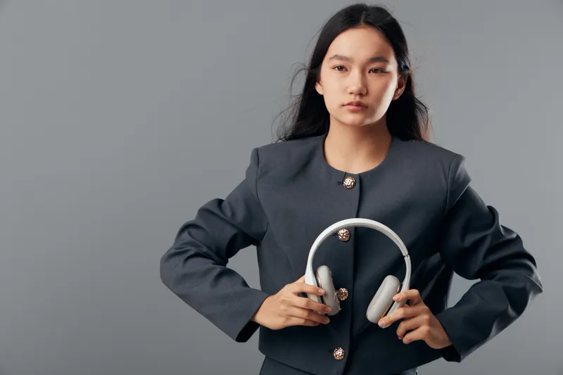 Young woman with long dark hair wearing modern gray jacket holding wireless headphones in studio. Female model posing with tech accessory for music and lifestyle concept on plain background.