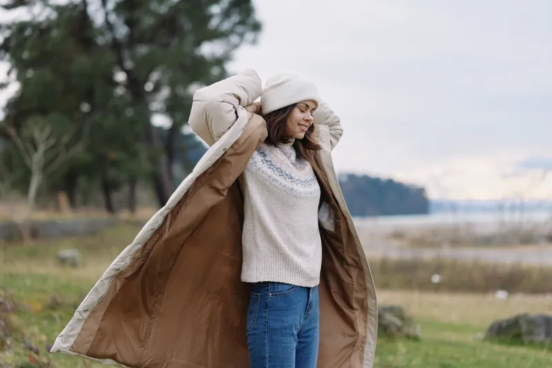 woman, coat, blanket, outdoor, autumn, cozy scene of a woman wrapping herself in a large coat beside a lake, wearing sweater and jeans in a chilly breeze