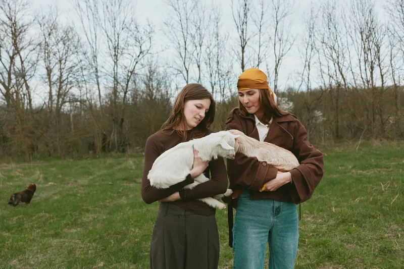 Two women outdoors cradle a white goat kid, sharing a gentle moment in a sunlit field, rustic jackets and vintage hats, capturing warmth, friendship, and a natural lifestyle in a pastoral countryside