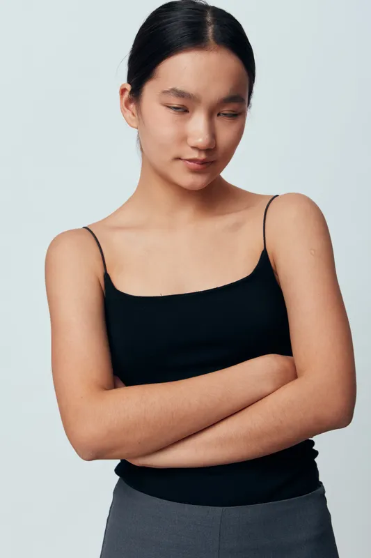 Young woman with closed eyes and calm expression standing with arms crossed wearing black tank top and gray pants against plain background in studio portrait.
