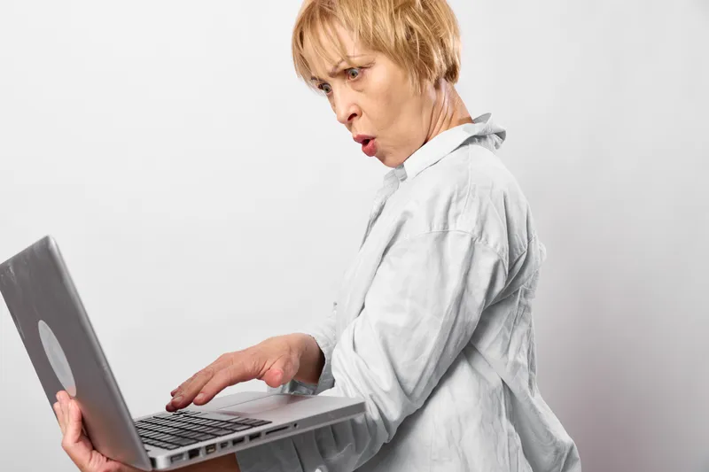 Elderly woman using laptop with surprised worried face, sitting indoors on white background, stress from technology online concern, close hands in frame