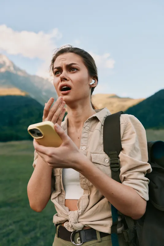 A woman outdoors with a backpack, holding a smartphone and reacting with surprise, standing in a mountain valley under a blue sky.