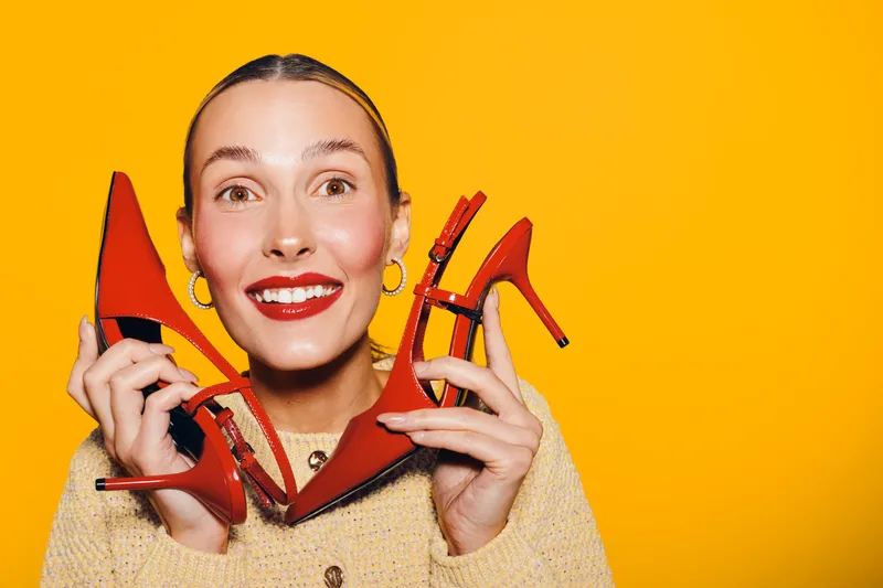 Happy woman with red lipstick holds red high heel shoes near face on bright yellow background. Concept of fashion, style, shopping, and joyful emotions in studio lighting.