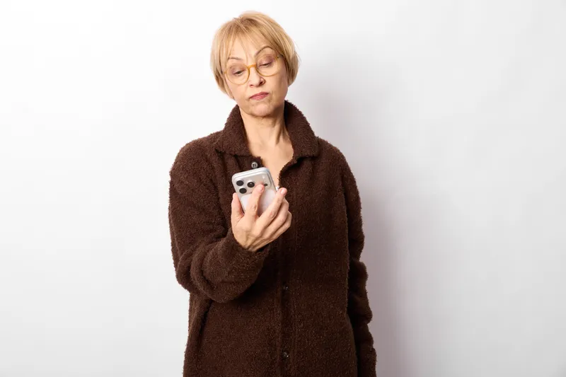 Elderly woman smartphone portrait indoors annoyed technology wireless moment, holding mobile device with both hands, frowning while viewing screen, white background