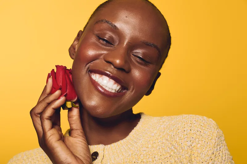Happy dark skinned African woman with closed eyes smiling holding red rose near face. Yellow background and cozy sweater create warm joyful mood. Concept for beauty and happiness.