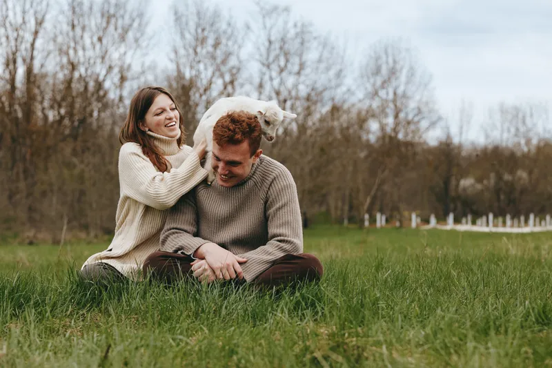 Young couple in cozy sweaters enjoys playful moment sitting on green grass in a natural outdoor setting during a cool day with bare trees in the background.