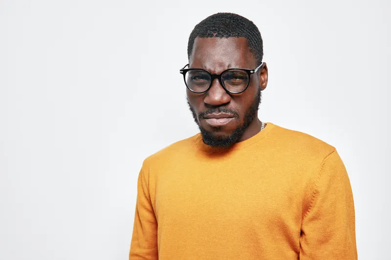 African man with dark skin wearing glasses and orange sweater standing against white background with serious expression and short hair in studio portrait.