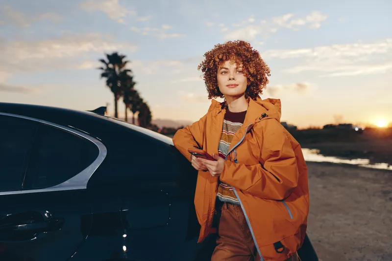 Woman lifestyle outdoors, sunset light, car exterior, orange coat, curly hair, portrait pose, street evening, calm mood, hands adjusting jacket, natural colors