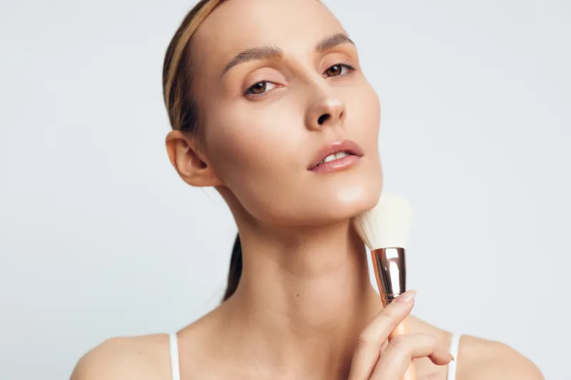 A young woman with clear skin holds a makeup brush near her neck, posing against a white background. The image represents skincare, beauty routine, and natural makeup application.