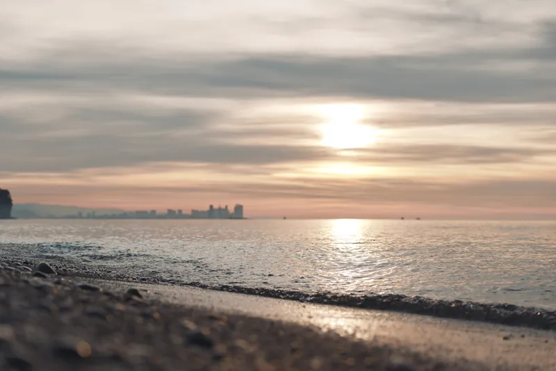 Sunset over a quiet beach with gentle waves, warm glow on the water and distant city skyline on the horizon, creating a serene coastal scene at dusk.