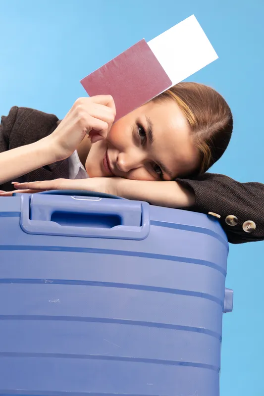 Young woman leans on blue suitcase holding passport and ticket on blue background. Travel concept with preparation, trip planning, and vacation mood in studio setting.