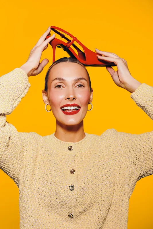 Smiling woman in beige sweater holds red high heel shoe above head on bright yellow background. Concept of fashion, style, happiness, and playful mood in studio portrait.