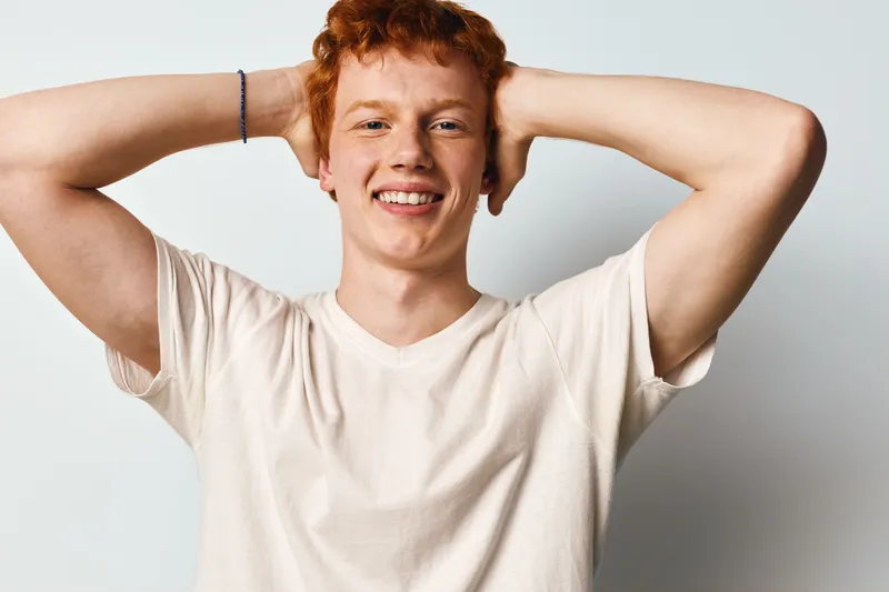 Young man with red hair and freckles smiles with hands behind head wearing white t shirt on light background. Casual male portrait with happy relaxed expression and natural look.
