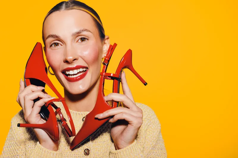 Smiling woman holds red high heels near face on yellow background. Fashion concept with bright colors, happy model, and stylish footwear for lifestyle and shopping themes.