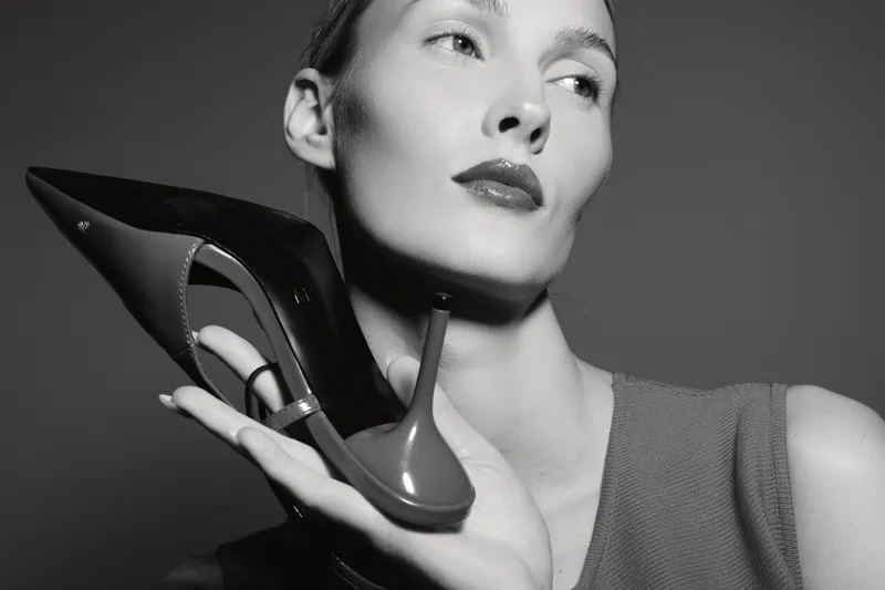 Portrait of a young white woman with fair skin holding a black high heel shoe near her face in monochrome.