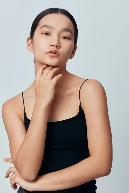 Young Asian woman in black tank top with natural makeup posing with hand on chin against light gray background. Beauty, confidence, fashion and portrait of female model in studio lighting.