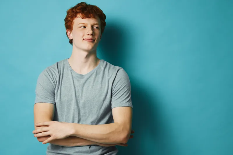 Young man with red hair wearing gray t shirt stands with arms crossed looking aside against blue background.