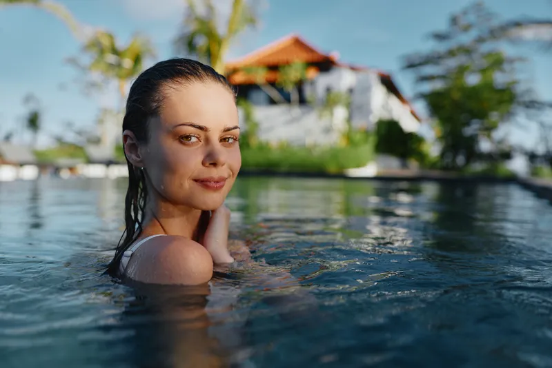 Smiling woman in a pool, wet hair, sunlit water and a tropical garden in the background, calm lifestyle moment, wellness and relaxation, soft natural light, warm mood.