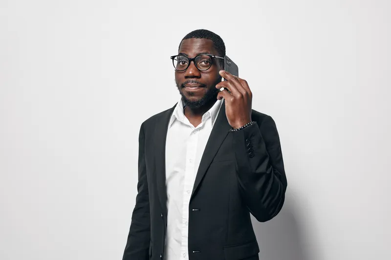 Black man with dark skin wearing glasses and black suit jacket talking on smartphone against white background. Professional African American businessman in formal outfit with confident expression.