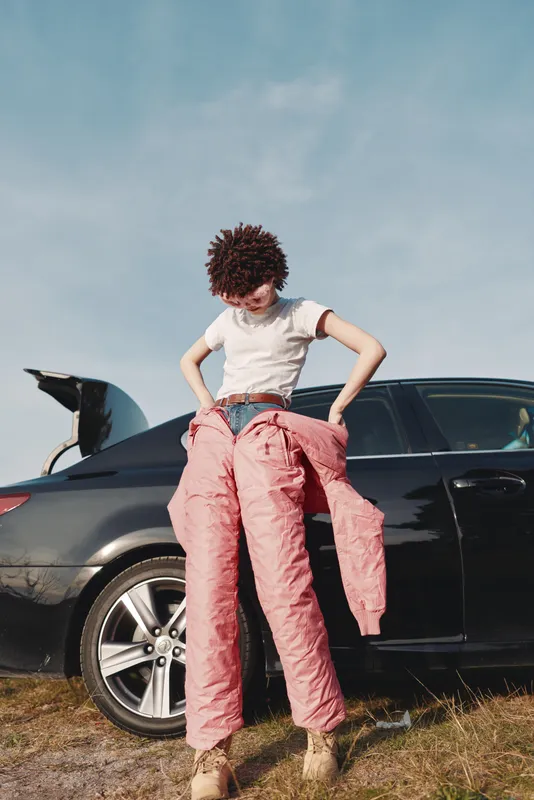 Fashion scene with an individual beside a sleek car, pulling on pink pants in a casual outdoor setting under a clear blue sky, showing a relaxed street style and confident pose.