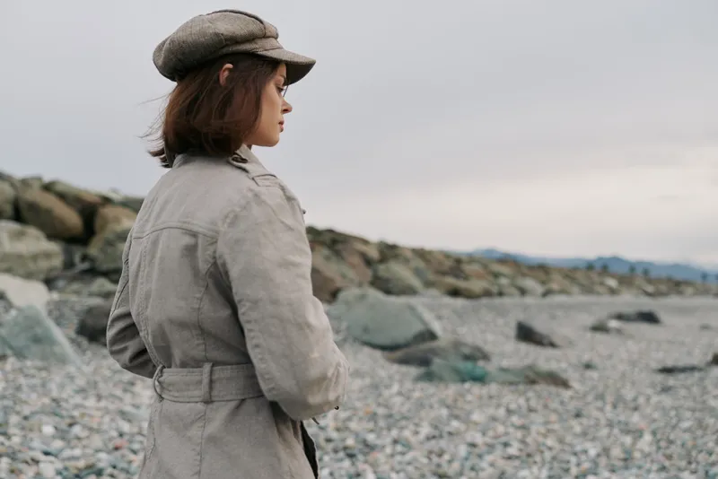 A young woman in a beige coat and hat stands on a rocky beach, looking thoughtfully into the distance. Overcast sky and calm atmosphere create a reflective mood.