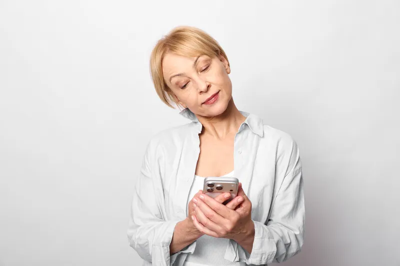 Woman smartphone portrait communication technology digital studio smiling female indoors, holding phone closeup, looking down softly, concept of messaging and online connection, neutral background