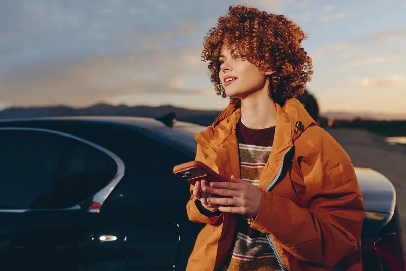 Woman with curly hair stands by a car outdoors at sunset, holding a smartphone and looking away. Lifestyle travel moment, warm evening light, casual jacket, relaxed mood today.