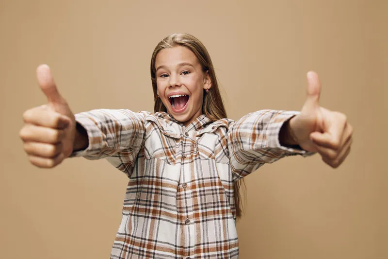 Happy girl with long hair smiling and showing both thumbs up wearing casual plaid shirt on beige background. Positive emotion, approval and cheerful child portrait.