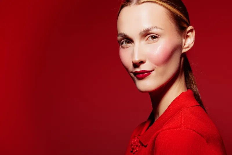 Portrait of confident woman with red lipstick and blush wearing red outfit on red background. Concept of fashion, beauty, makeup, and elegance in studio lighting.