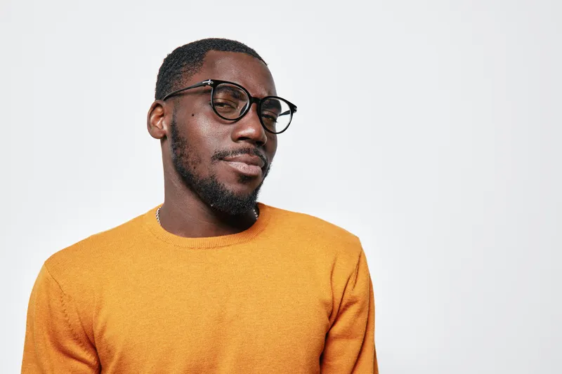 Portrait of confident African American man with dark skin wearing glasses and orange sweater looking sideways on white background. Casual style and thoughtful expression in studio.