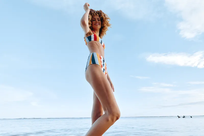 Woman in bikini on beach by ocean in summer, joyful carefree mood, curly hair and raised arm, sunny outdoor scene, blue sky and calm water, lifestyle vacation concept, for relaxation