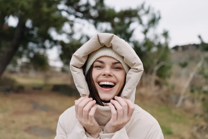 Woman outdoors in a hooded coat, smiling with joy in a natural landscape, casual lifestyle portrait showcasing warmth and carefree mood