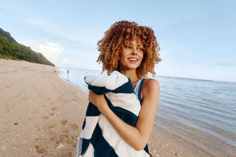Young woman with curly hair wrapped in a striped towel smiles while standing on a sandy beach near the calm sea under a clear sky.